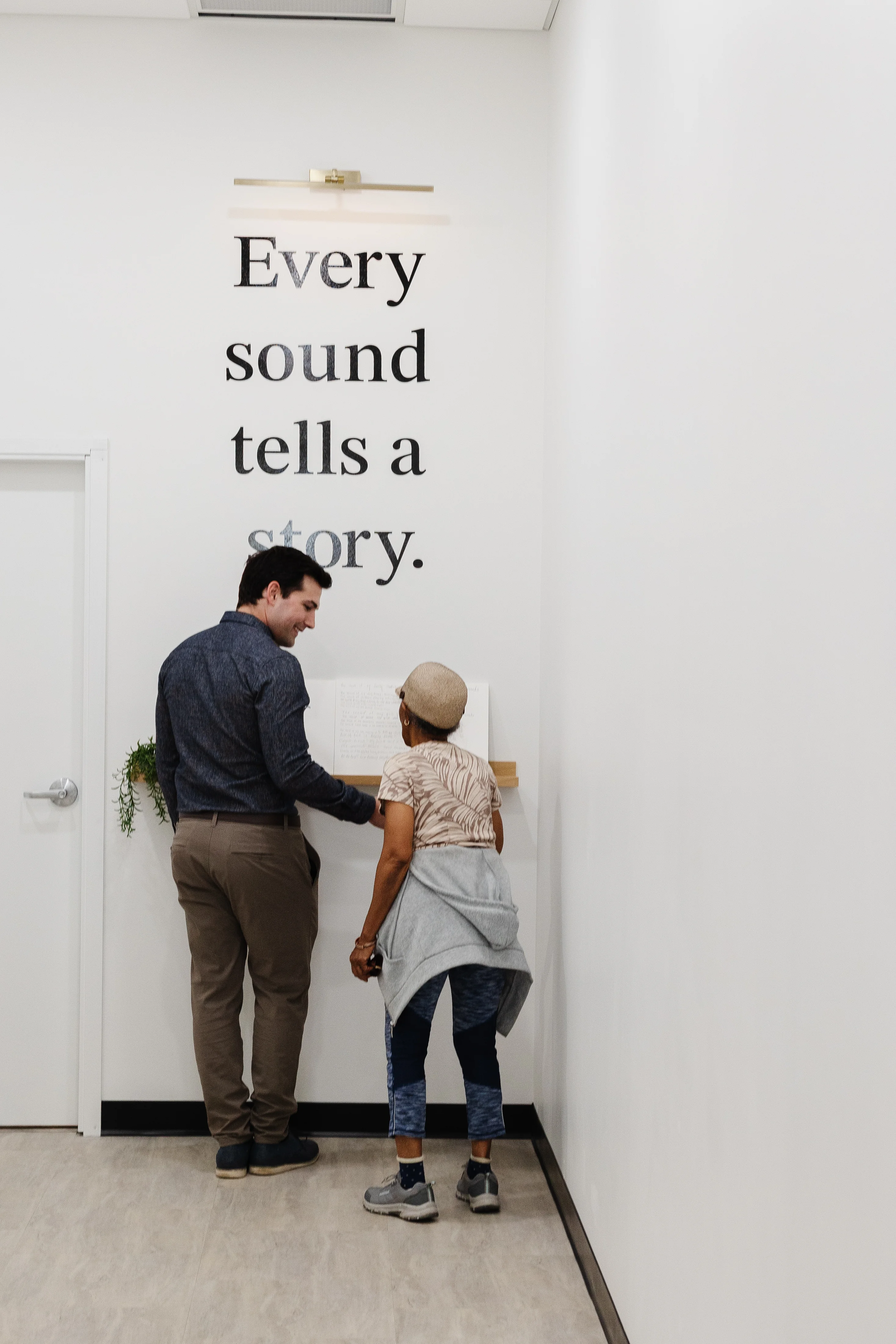 Armando Di Lello with a patient at simplyHEAR in front of the 'Every sound tells a story' wall
